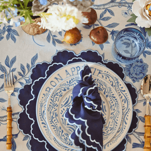 Decorative table setting with a blue and white plate, napkin, and cutlery on a floral tablecloth. Malmaison - Luxe & Beau - Versailles Embroidery Heirloom Placemats - Navy