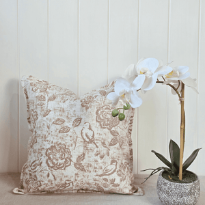 Decorative linen square cushion with floral and bird pattern next to a potted orchid on a neutral background