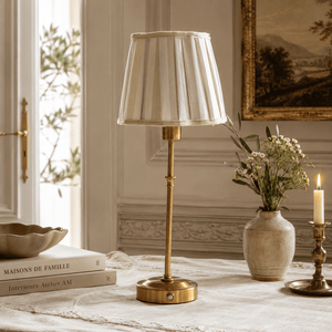A table lamp with a pleated cream fabric shade and a polished brass base stands on a draped tablecloth in a styled interior setting. A vase with flowers, a candle, books, and wall molding are visible around it. Touch lamp with beige shade and gold base on French Country styled table