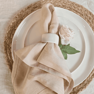 Blush linen napkin styled on white ceramic plate and rose with simple table setting on a woven placemat