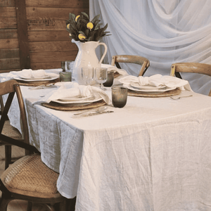 Elegant table setting with white French Linen tablecloth, plates, glasses, and a vase with flowers in a rustic indoor setting.