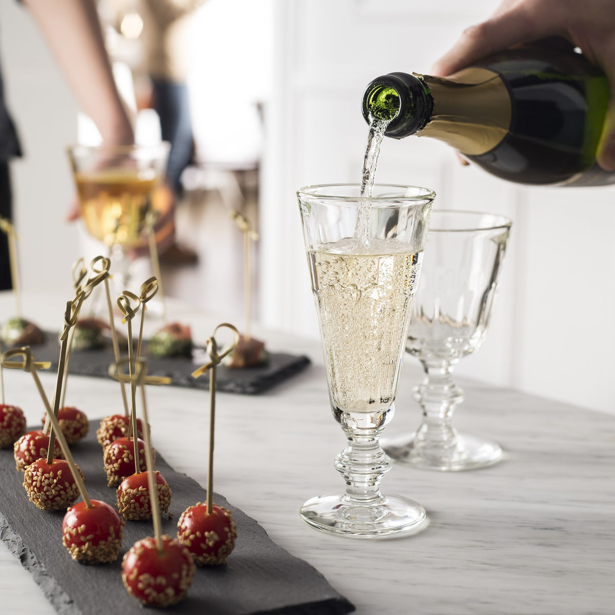 Person pouring champagne into a La Rochere Perigord Champagne glass with appetizers on a table.