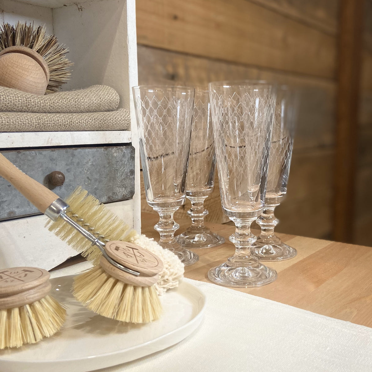 A set of four etched crystal champagne flutes with fishnet patterns, displayed alongside a wooden storage box and a broom, on a table with a floral fabric.