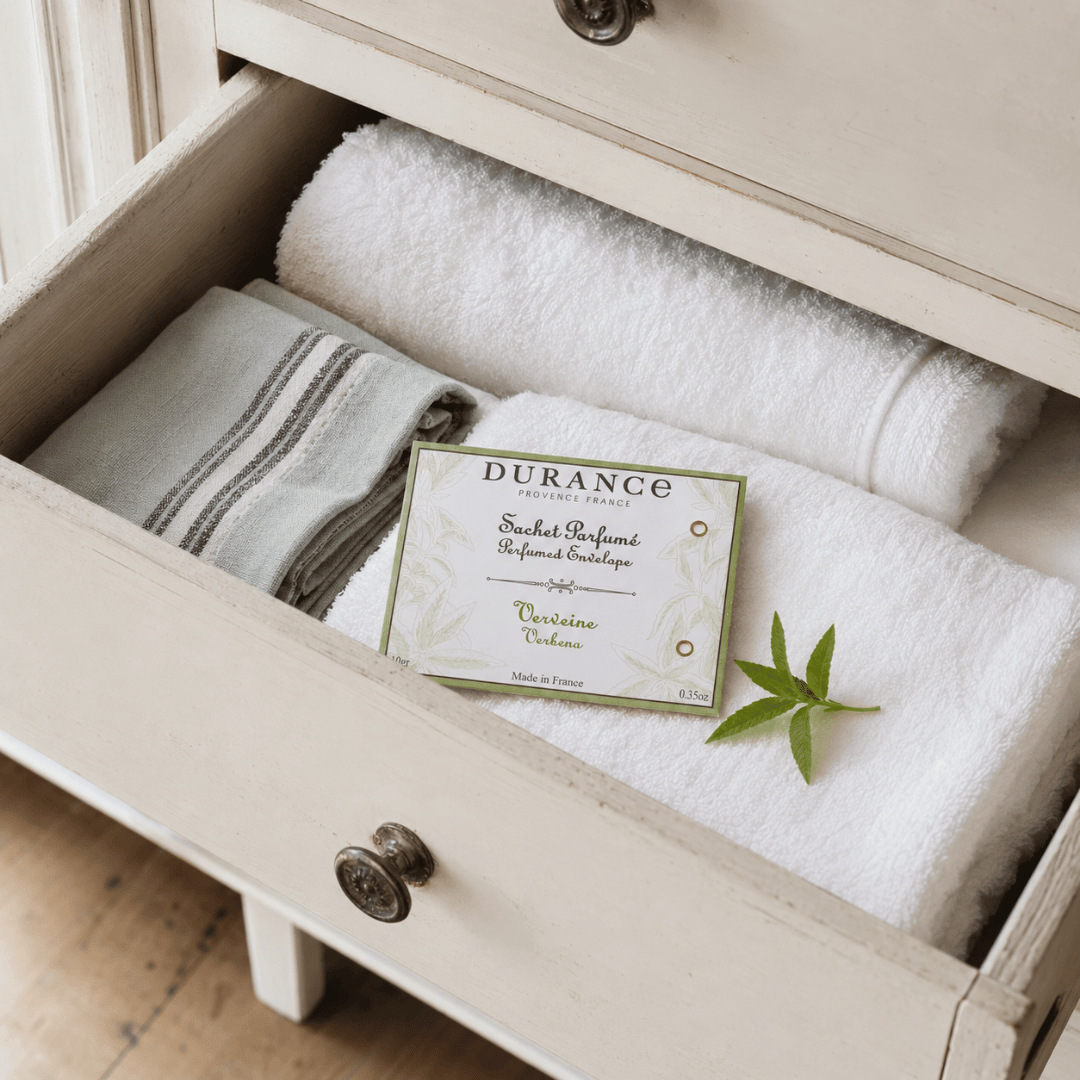 A Durance Scented envelope nestled inside an open, distressed cream-colored wooden drawer, surrounded by folded white towels and a small green Verbena leaf.