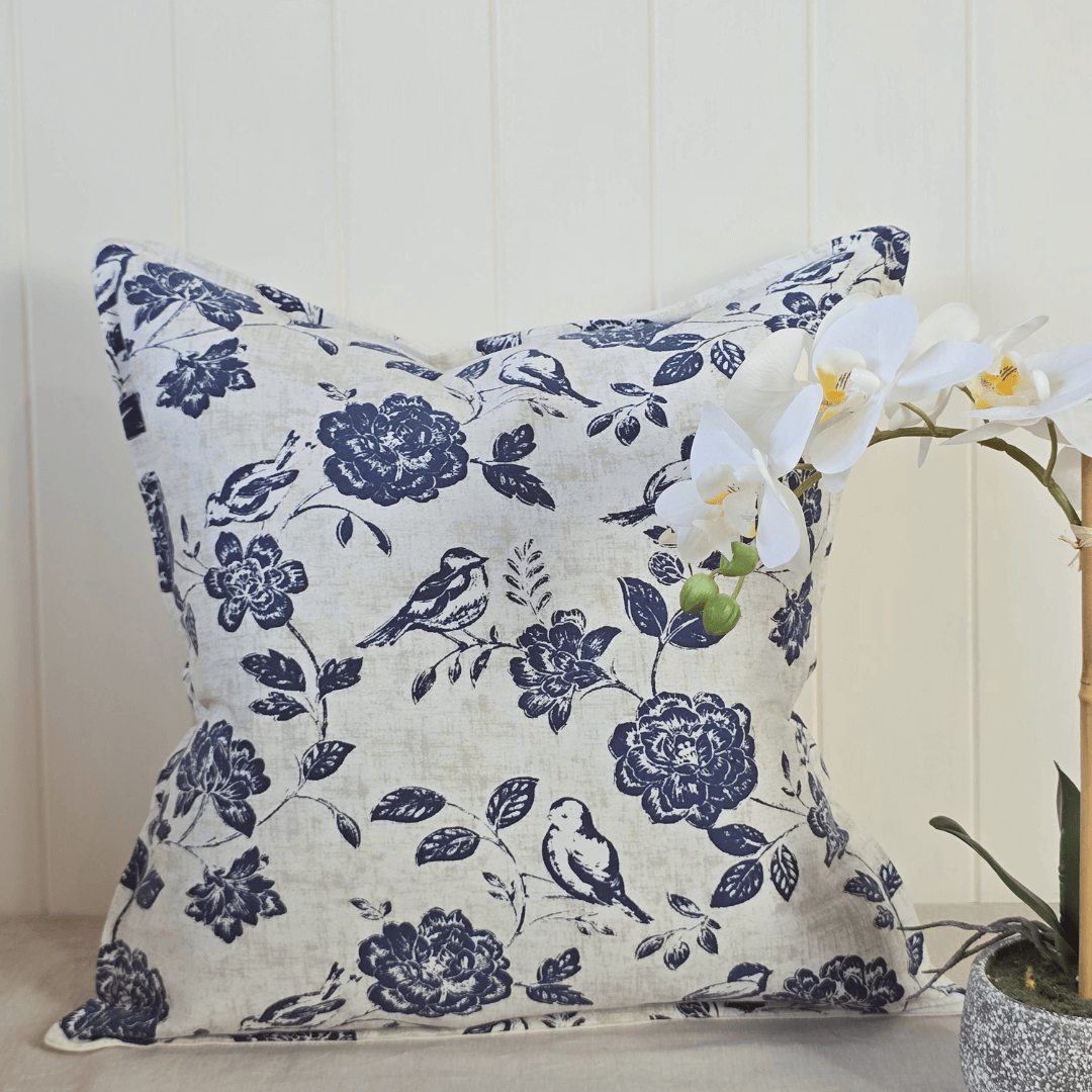 Decorative square cushion with floral and bird pattern on a neutral background and orchid in pot