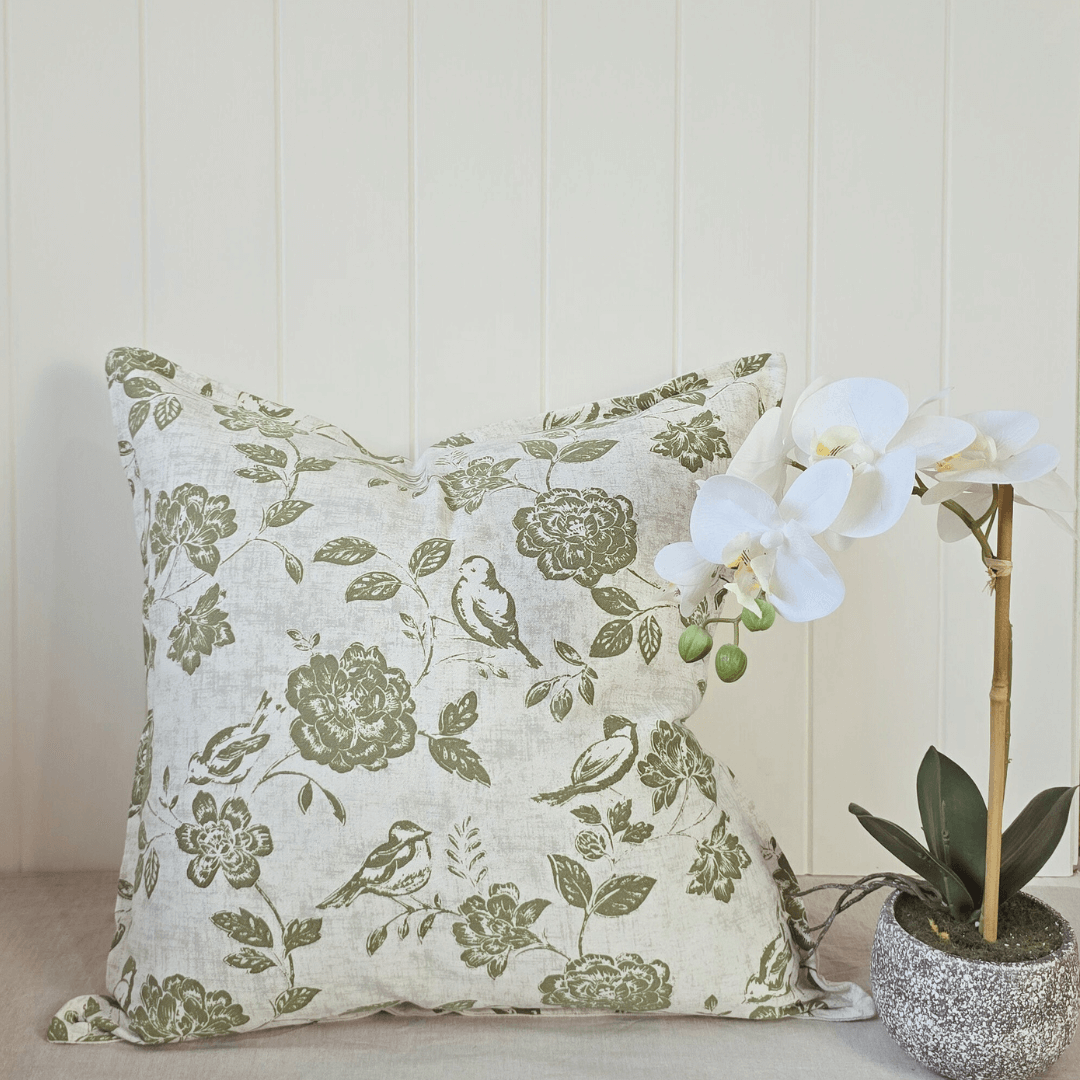Decorative square cushion with floral and bird pattern next to a potted orchid on a light wooden surface.