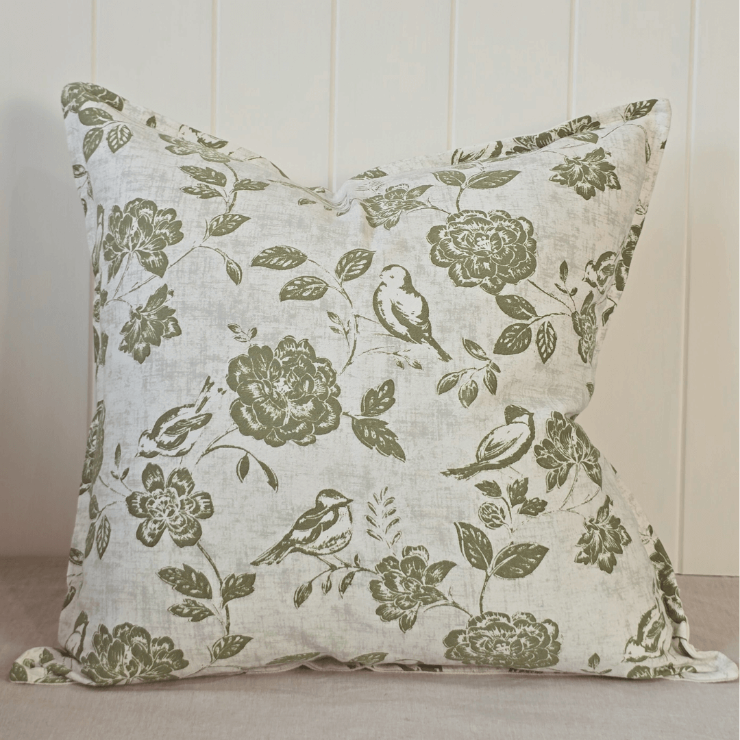 Decorative square cushion with floral and bird pattern next to a potted orchid on a light wooden surface.