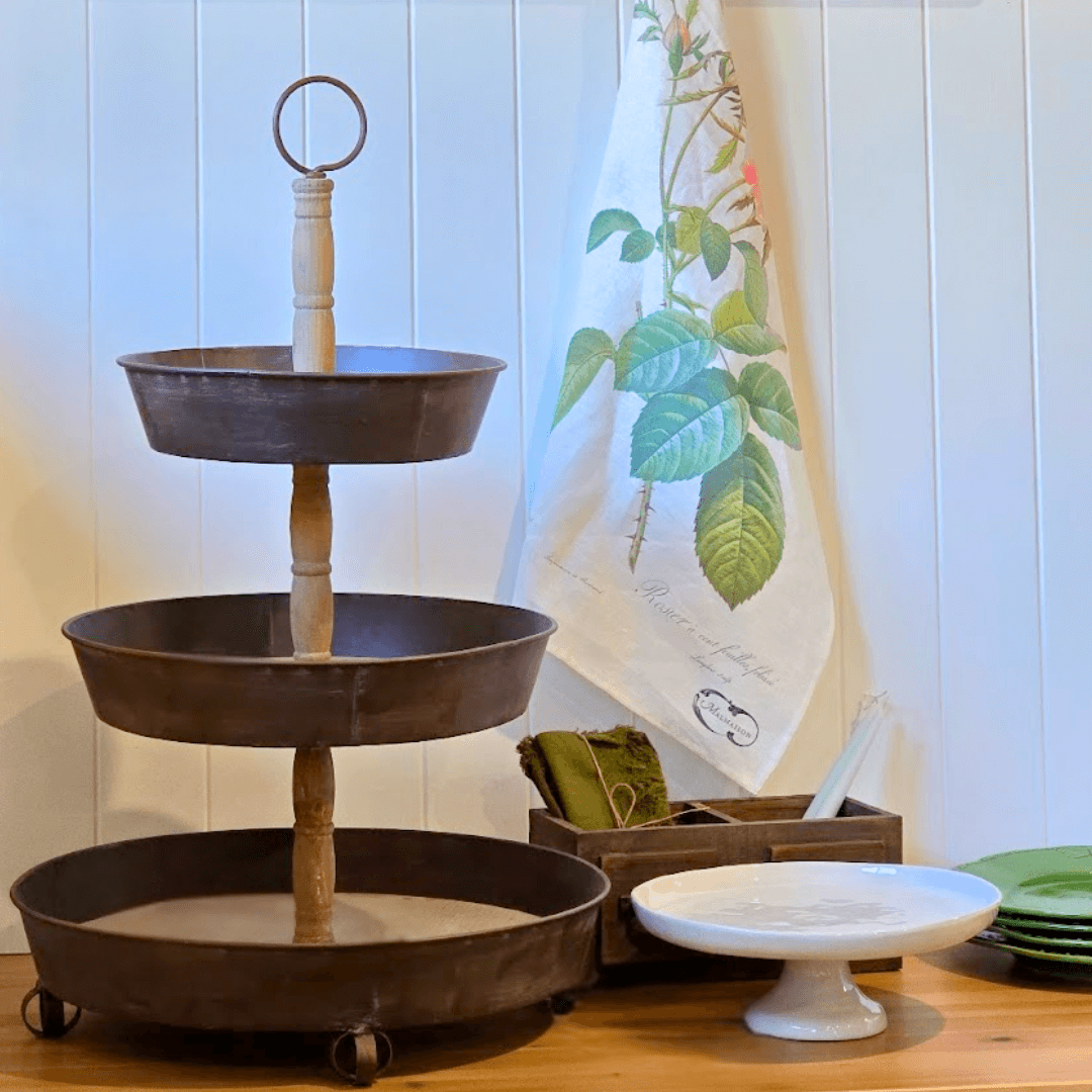 Three-tiered French Country metal stand on a wooden surface with a decorative towel in the background.