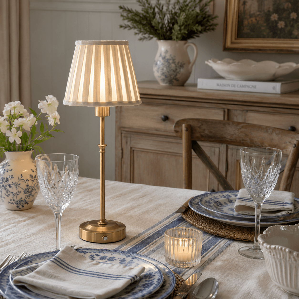A table lamp with a pleated cream fabric shade and a polished brass base stands on a draped tablecloth in a styled interior setting. A vase with flowers, a candle, books, and wall molding are visible around it.  Touch lamp with beige shade and gold base on French Country styled table