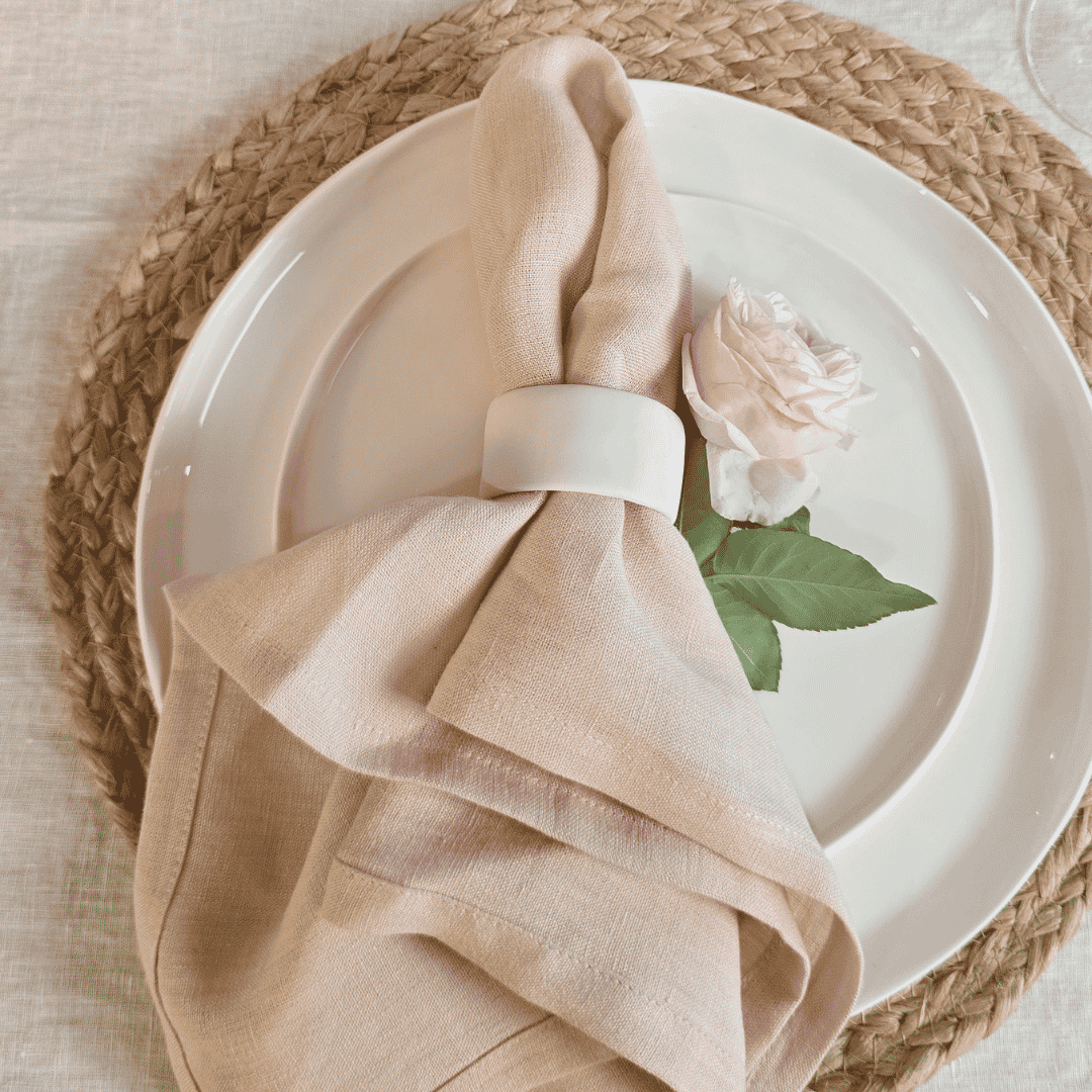 Blush linen napkin styled on white ceramic plate  and rose with simple table setting on a woven placemat