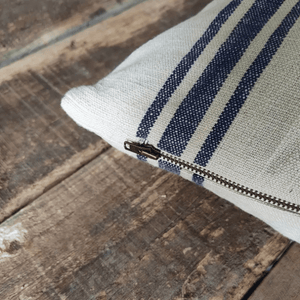 A close-up of a French grain stripe cushion in a natural linen-like fabric with dark navy blue stripes, featuring a brass zipper.