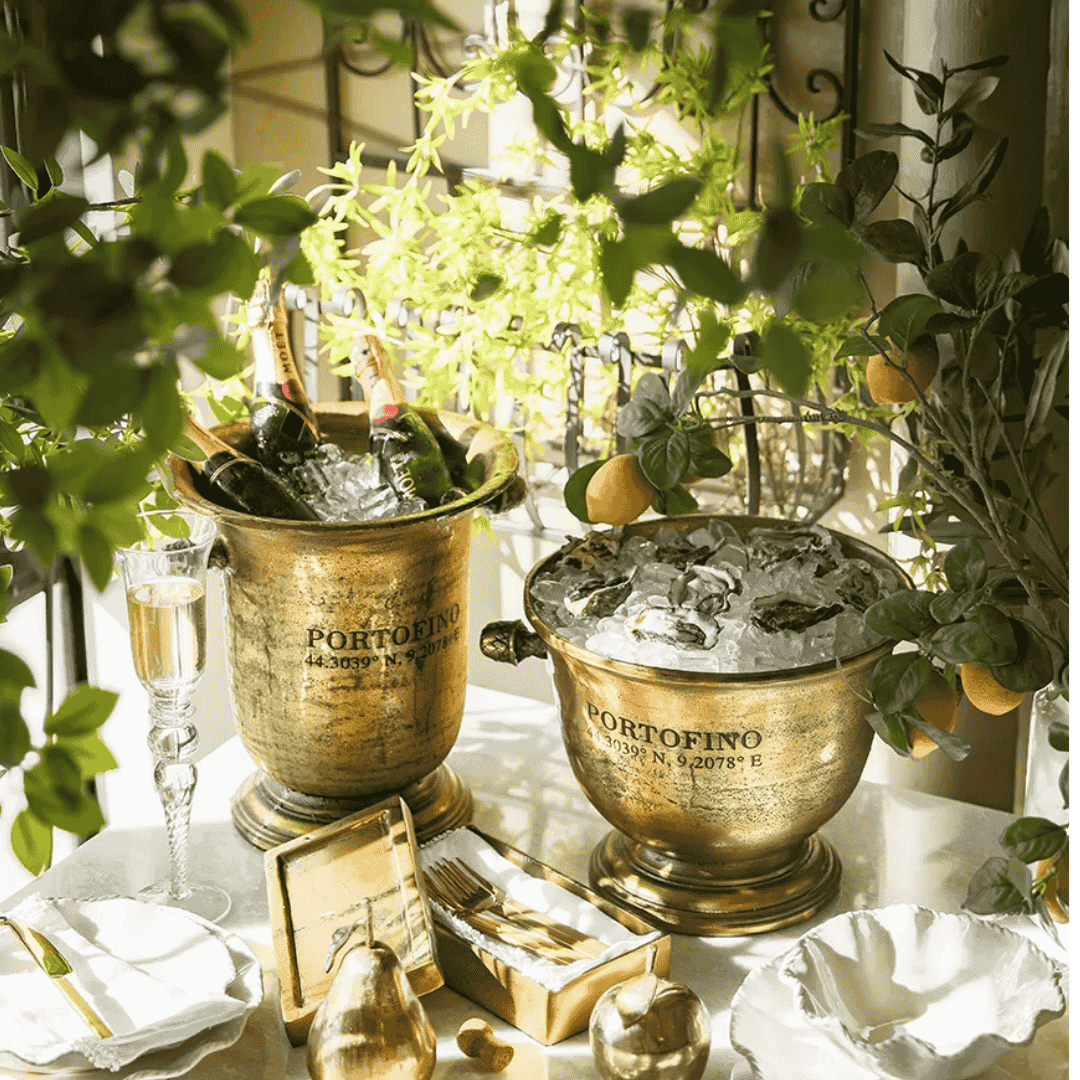 Gold Portofino ice buckets with champagne and fruit on a table with greenery.