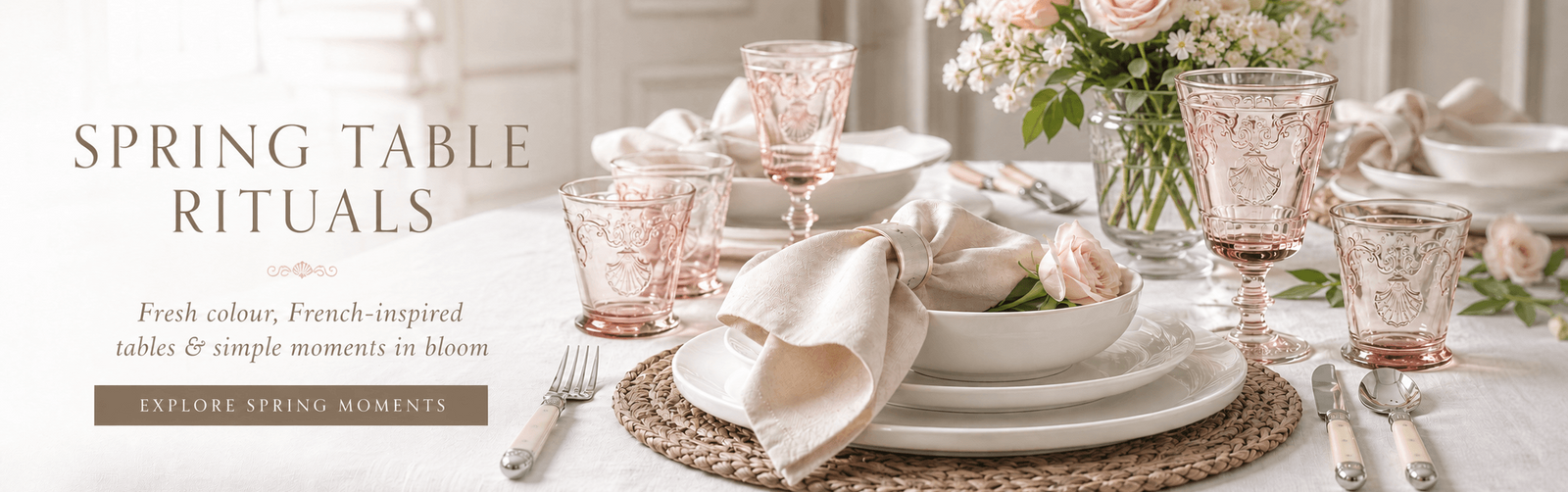 A French styled spring table setting with white plates, beige napkins, pink French glassware, woven placemats, and a floral centerpiece on a white tablecloth. 