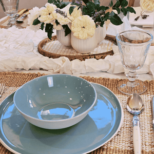 Table setting with blue plates, a bowl, and silverware on a woven Lucien jute placemat. Rectangle.