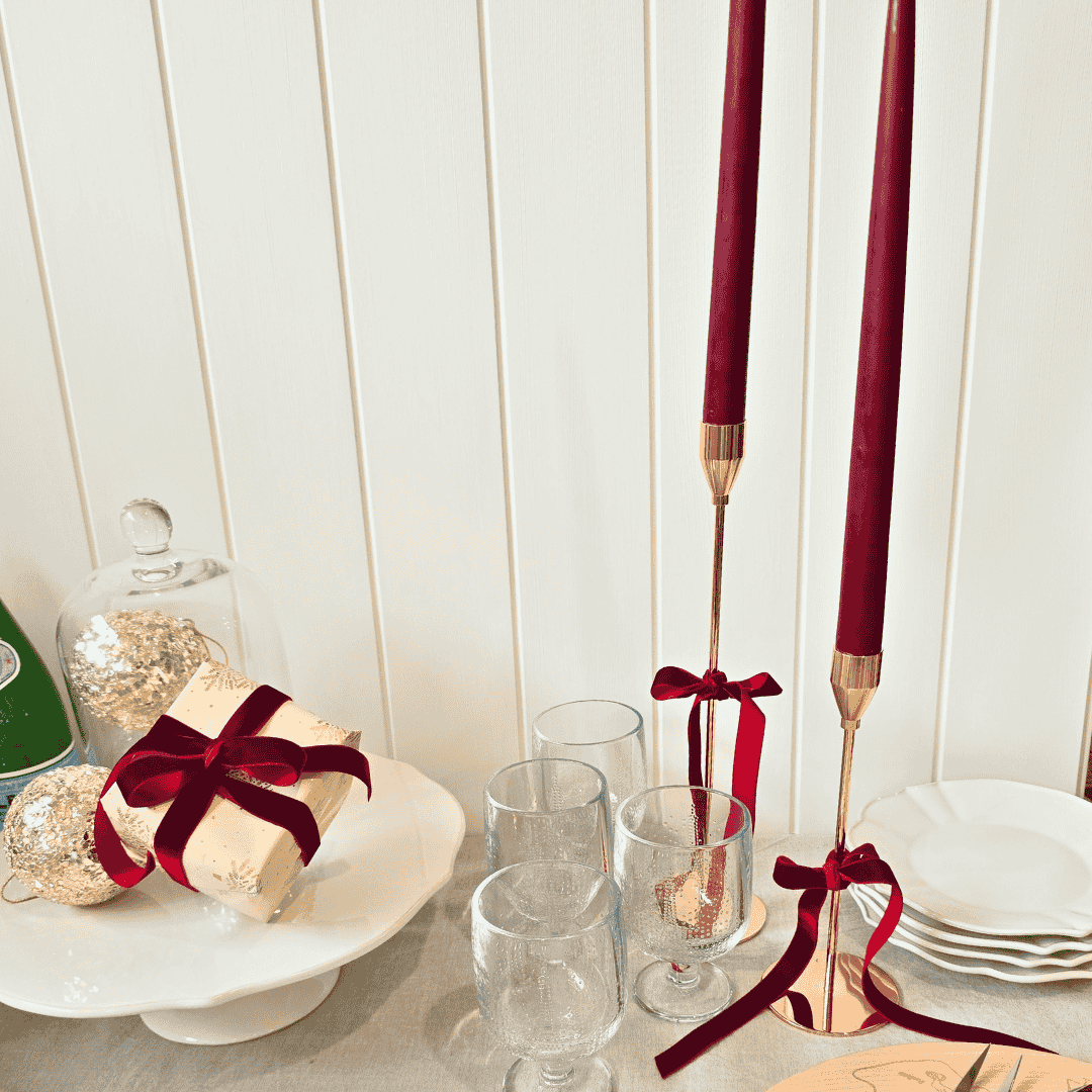 Decorative table setting with red taper candles, gift box, and glasses on a white surface.